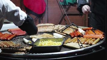 salsichas grelhadas e batatas fritas em uma grande prateleira de metal no festivo mercado noturno. comida de rua. feiras de natal e ano novo - vendedores de balcões de fast food. video