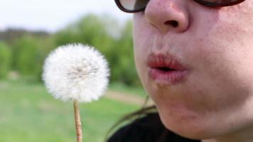 Portrait of a beautiful young woman on a summer lawn blowing on a ripe dandelion on a sunny day outdoors. Enjoy nature. Allergy free concept.