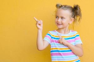 la niña sonriente se siente atraída por la atención que señala con el dedo el espacio de la copia. el concepto de publicidad de productos, bienes y servicios. aislado sobre fondo amarillo foto