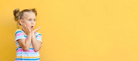 Little child girl in striped colored summer t-shirt surprised expression looks at copy space on yellow background, studio portrait.Advertising of children's products and sale. Banner photo
