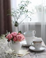 un jarrón de rosas rosadas y tazas blancas sobre una mesa junto a la ventana. primavera verano naturaleza muerta foto