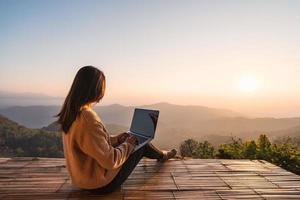 joven viajera independiente que trabaja en línea usando una computadora portátil y disfrutando del hermoso paisaje natural con vista a la montaña al amanecer foto