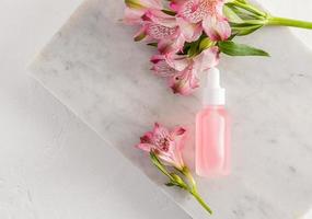nature cosmetics in a glass matte bottle with a dropper and pink spring flowers against the background of marbled white tiles. self-care.