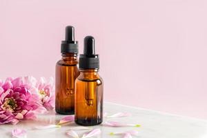 two cosmetic bottles with a pipette with serum for skin care of the face and body stand on a marble podium with flowers. pink background. copy space. photo