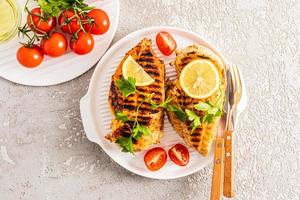 two juicy grilled chicken breasts on a white ceramic tray for serving with vegetables and lemon. top view. cement grey background. delicious food.