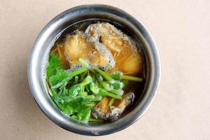 Top view of fried fish and chinese celery, young ginger slice in broth, Chines style soup in round stainless bowl on light brown wood. photo