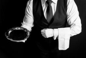 retrato de mayordomo o camarero con chaleco negro y guantes blancos sosteniendo una bandeja de plata con una servilleta sobre el brazo. foto
