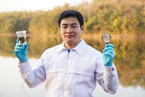 Asian man ecologist holds magnifying glass to inspect water in transparent glass from the lake. Concept, explore, analysis water quality and creature from natural source. Ecology field research. photo