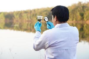 Back view of Asian man ecologist hold magnifying glass to inspect water in transparent glass from the lake. Concept, explore, analysis water quality and creature from natural source. Ecology field photo