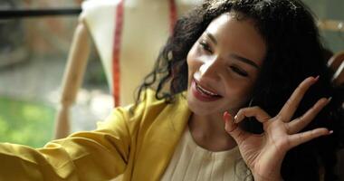 toma de mano, cara de una atractiva joven diseñadora sonriendo y saludando mientras usa una videollamada de teléfono inteligente durante la venta en línea en una sastrería, una pequeña empresa de inicio video