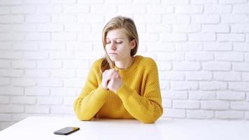 mujer pensativa con suéter amarillo sentada en la mesa pensativa, usando un teléfono inteligente video