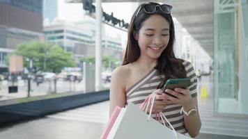 retrato de moda de estilo de vida feminino asiático feliz moderno. linda jovem atraente desfruta de um passeio elegante com pacotes de sacolas de compras usa smartphone na rua, roupa da moda no shopping video