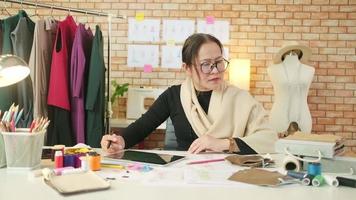 Aziatisch middelbare leeftijd vrouw mode ontwerper werken in studio door idee tekening schetsen met digitaal tablet en kleurrijk kleding stof voor een jurk ontwerp verzameling, professioneel winkel kleermaker mkb ondernemer. video