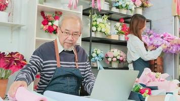 el anciano asiático propietario de una floristería vive en línea transmitiendo para vender en un sitio web de comercio electrónico, empleado y cliente con flores detrás, trabajando en una floristería colorida, un pequeño empresario.