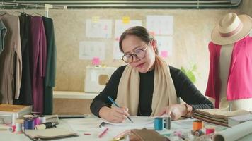 asiatisk medelålders kvinna mode designer arbetssätt i en studio förbi aning teckning skisser med färgrik tråd och sömnad för en klänning design samling, professionell boutique skräddare sme entreprenör. video