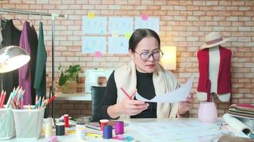 asiatische modedesignerin mittleren alters arbeitet in einem kreativstudio, skizziert, trinkt kaffee, denkt an ideen, stellt kleiderdesign-kollektionen dar und ist eine professionelle boutique-schneiderin und kleine unternehmerin. video