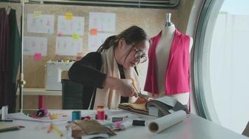 Asian middle-aged female fashion designer works in studio, cutting and choosing fabric and thread colors with drawing sketches for dress design collections. Professional boutique tailor entrepreneur. video