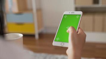 Closeup of hand woman using smartphone with green screen while sitting in living room. Blank digital smartphone in hand girl. Showing content videos blogs tapping on center screen.