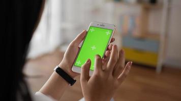 Closeup of hand woman using smartphone with green screen while sitting in living room. Blank digital smartphone in hand girl. Showing content videos blogs tapping on center screen.