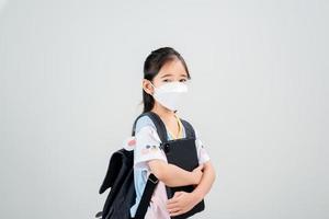 niña asiática lleva una mochila con máscara de respirador para proteger el brote de coronavirus y apunta con la mano al fondo en blanco, nuevo virus covid-19 foto