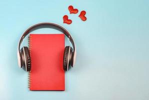 flat lay of red notebook covered with headphones on blue background decorated with red glitter hearts , copy space. Audio book, podcast, .love diary, valentine's day. photo