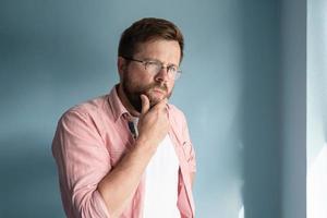 Caucasian man in glasses looks incredulously, suspiciously with a squint, thinking about something and touches his beard with hand. photo