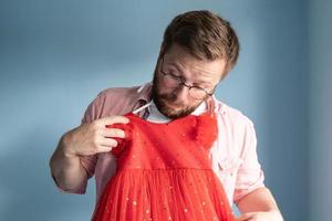 Funny father holds a beautiful red dress of his daughter in hands, jokingly tries it on himself and looks thoughtfully. photo