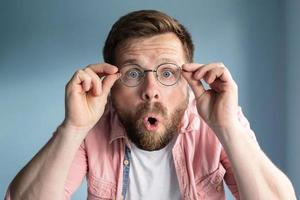 Man is shocked, he does not believe his eyes, he holds glasses with hands and opens mouth in surprise. Blue background. photo