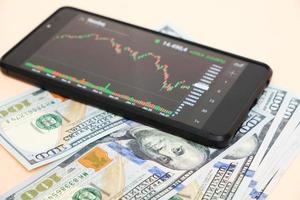 Stock exchange chart on smartphone screen and hundred dollar bills on beige background.