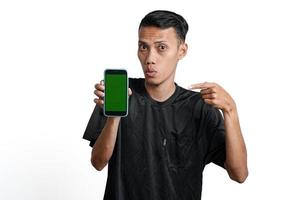 emocionado hombre asiático con camiseta negra de entrenamiento, apuntando a la pantalla verde del teléfono inteligente. aislado por fondo blanco foto