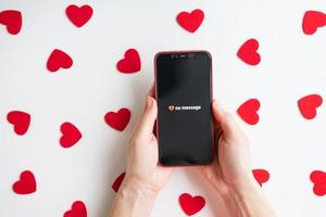 On a white table against the background of small red hearts, a girl is holding a smartphone with an activated screen and no SMS message in the form of a broken heart. photo