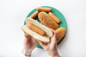 Fresh buns for making hot dogs that lie on a green plate along with sausages that the girl holds in her hands. Top view, close-up. photo