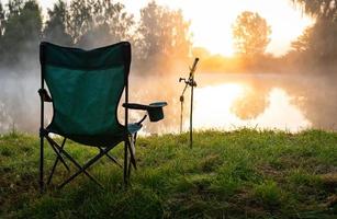 silla de pescador y cañas de pescar en el fondo sobre el lago al amanecer, mañana nublada. concepto de pesca. foto