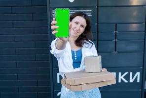 mujer feliz sosteniendo un teléfono móvil con una maqueta de pantalla verde para la inscripción y sosteniendo muchas cajas de cartón diferentes con pedidos en línea, satisfecha con la entrega rápida, comprando productos de alta calidad. foto