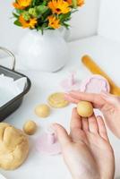 preparando la masa para hacer galletas con moldes en forma de flores, un rodillo, un ramo de flores naranjas y una bandeja para hornear, la niña forma galletas en sus manos. concepto de cocina familiar foto