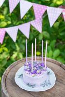 Very beautiful bento cake with purple, veri peri, matthiola flowers with green leaves, candles in the cake on the background of pink flags. Birthday. Vertical photo. photo