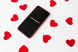 A smartphone with an activated screen with an SMS message in the form of a heart lies on a white table against a background of small red hearts. Business technology and social lifestyle concept. photo