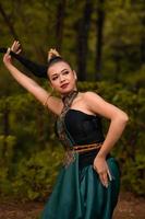 Asian woman holding her black hair in a green costume while wearing makeup and posing in front of the forest during the dance festival photo