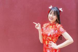 linda chica asiática con el pelo largo que usa un vestido rojo cheongsam en el tema del año nuevo chino mientras muestra su mano para presentar algo sobre un fondo rojo. foto