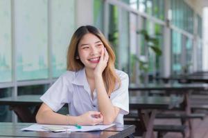 una joven estudiante asiática linda está usando un teléfono inteligente para buscar información para un informe de estudio que sonrió alegremente mientras estaba sentada en la universidad. foto