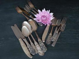 Tarnished silverware on a dark background with a single pink flower. photo