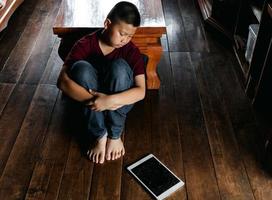 retrato de un niño asiático, cara triste de niño, niño infeliz sentado abrazando las rodillas miró la tableta rota en el suelo, emoción cara triste y triste, concepto de niños deprimidos foto