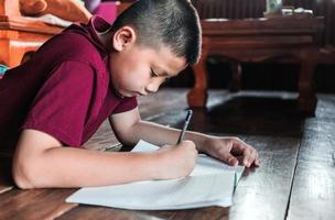 primer plano de un niño asiático sentado en el suelo de madera escribiendo un libro o haciendo deberes con una cara muy determinada en casa. foto