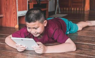 Little smiling Asian boy sitting on the wooden floor and playing on a digital tablet at home. Kid in his using smart devices having fun online with his friends in a video call photo