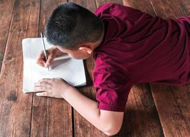 niño sentado en el suelo de madera escribiendo un libro o haciendo la tarea en casa. vista superior del libro de escritura para niños foto