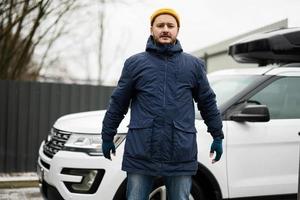 Man driver wear jacket and yellow hat against his american SUV car with roof rack in cold weather. photo