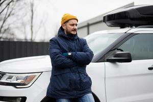 Man driver wear jacket and yellow hat against his american SUV car with roof rack in cold weather. photo