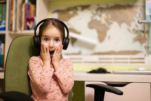 Young girl wearing headphones, enjoys her remote education. Distant studying, homeschooling. Funny face surprised. photo