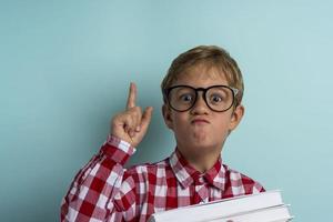 un niño con gafas levanta el dedo índice, libros en las manos, sobre un fondo turquesa. el conocimiento es poder foto