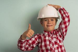 A cute boy in a plaid shirt and a white construction helmet holds a thumbs up, a portrait of a small builder photo